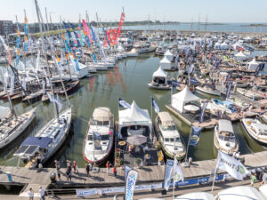 Overzicht van een drukbezocht haventerrein tijdens een botenbeurs met talloze zeil- en motorboten aan steigers en promotiekramen langs de kade.