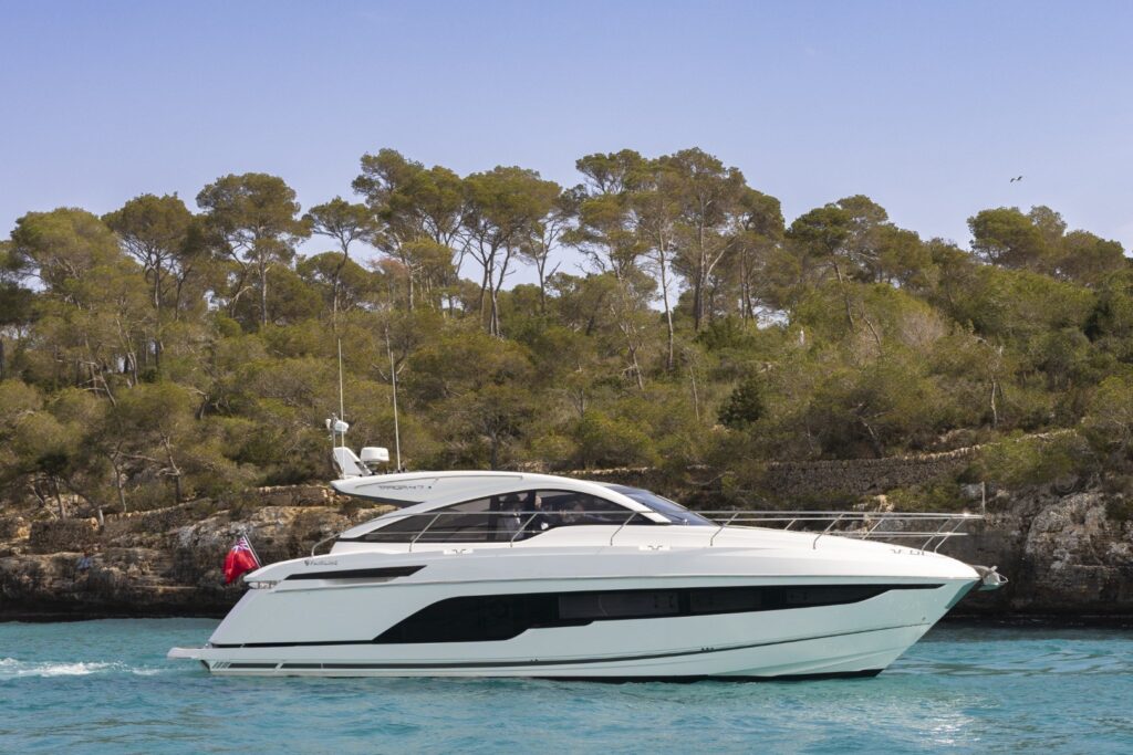 Een luxueuze Fairline Targa 47 motorboot vaart langs een groene kustlijn met bomen onder een heldere blauwe hemel.